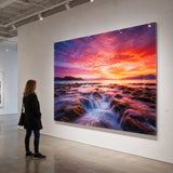Acrylglas bedrucken als großformatiges Wandbild in einer Kunstgalerie. Das Bild zeigt eine beeindruckende Landschaftsaufnahme eines Sonnenuntergangs über dem Meer. Durch den Direktdruck auf Acrylglas entstehen eine enorme Farbtiefe und eine glänzende Oberfläche, die eine Betrachterin im Raum bewundert.