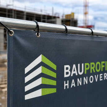 Bauzaunbanner – eine Detailaufnahme, die zeigt, wie ein bedrucktes Werbebanner mit schwarzen Kabelbindern durch Metallösen sicher an den Rohren eines Bauzauns befestigt ist.