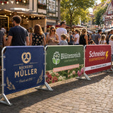 Hussen für Absperrgitter – verschiedene farbenfrohe Hussen für lokale Unternehmen wie Bäckereien und Gartencenter, die bei einem belebten Stadtfest an Absperrgittern befestigt sind.