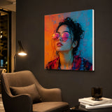 Leinwand Bedrucken – ein modernes, farbenfrohes Porträt einer jungen Frau mit runder Sonnenbrille in intensivem orange-blauem Neonlicht, das als Wandbild an einer dunklen Wand über einem grauen Sessel hängt.