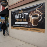 PVC Werbebanner der „River City Coffee Co.“, das direkt an einer Außenhauswand befestigt ist. Das Banner zeigt Kaffeebohnen und eine Tasse Kaffee und dient als hochwertige Außenwerbung für das lokal ansässige Café. Die Befestigung erfolgt über Metallösen am Rand des Banners.