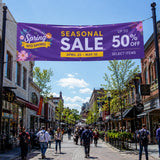 PVC Werbebanner in leuchtendem Lila mit der Aufschrift „Seasonal Sale“, das über eine belebte Einkaufsstraße gespannt ist. Das Banner ist wetterfest, verfügt über Metallösen zur Aufhängung und wirbt für eine Frühlingsaktion mit bis zu 50 % Rabatt.