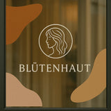Transparente Fensteraufkleber mit Frauen-Logo und Schriftzug Blütenhaut auf einer Salon-Glasscheibe.