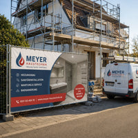Bauzaunbanner – ein individuell bedrucktes Werbebanner für einen Sanitär- und Heizungsbetrieb, das an einem Bauzaun vor einem eingerüsteten Wohnhaus befestigt ist und die Leistungen des Handwerkers sowie ein Beispielfoto eines Badezimmers zeigt.