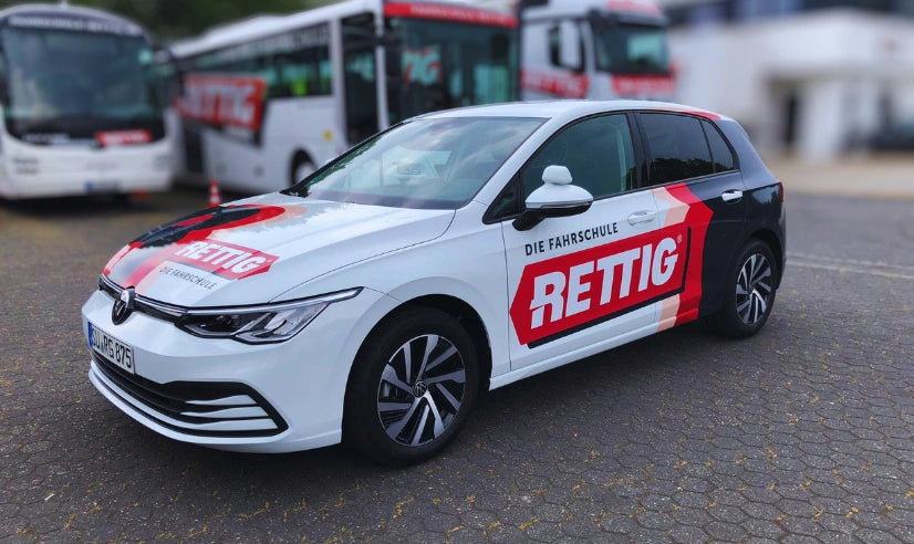 Fahrzeugbeschriftung einer Fahrschule Fahrzeugflotte mit beschriftetem Fahrschulauto sowie Fahrschulbus und LKW der Fahrschule Rettig im Hintergrund.
