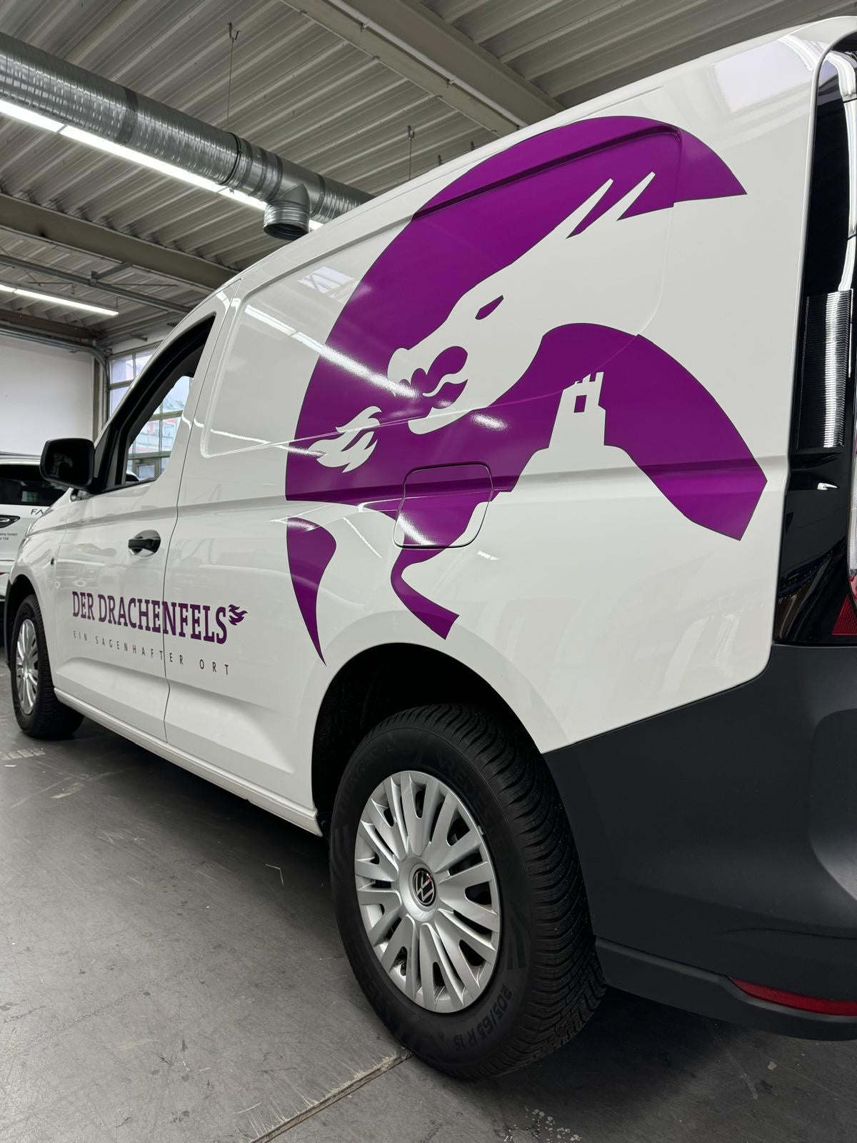 Fahrzeugbeschriftung mit Teilfolierung eines weißen Transporters mit großem lila Drachenmotiv und Firmenlogo „Der Drachenfels“ auf der Fahrzeugseite.