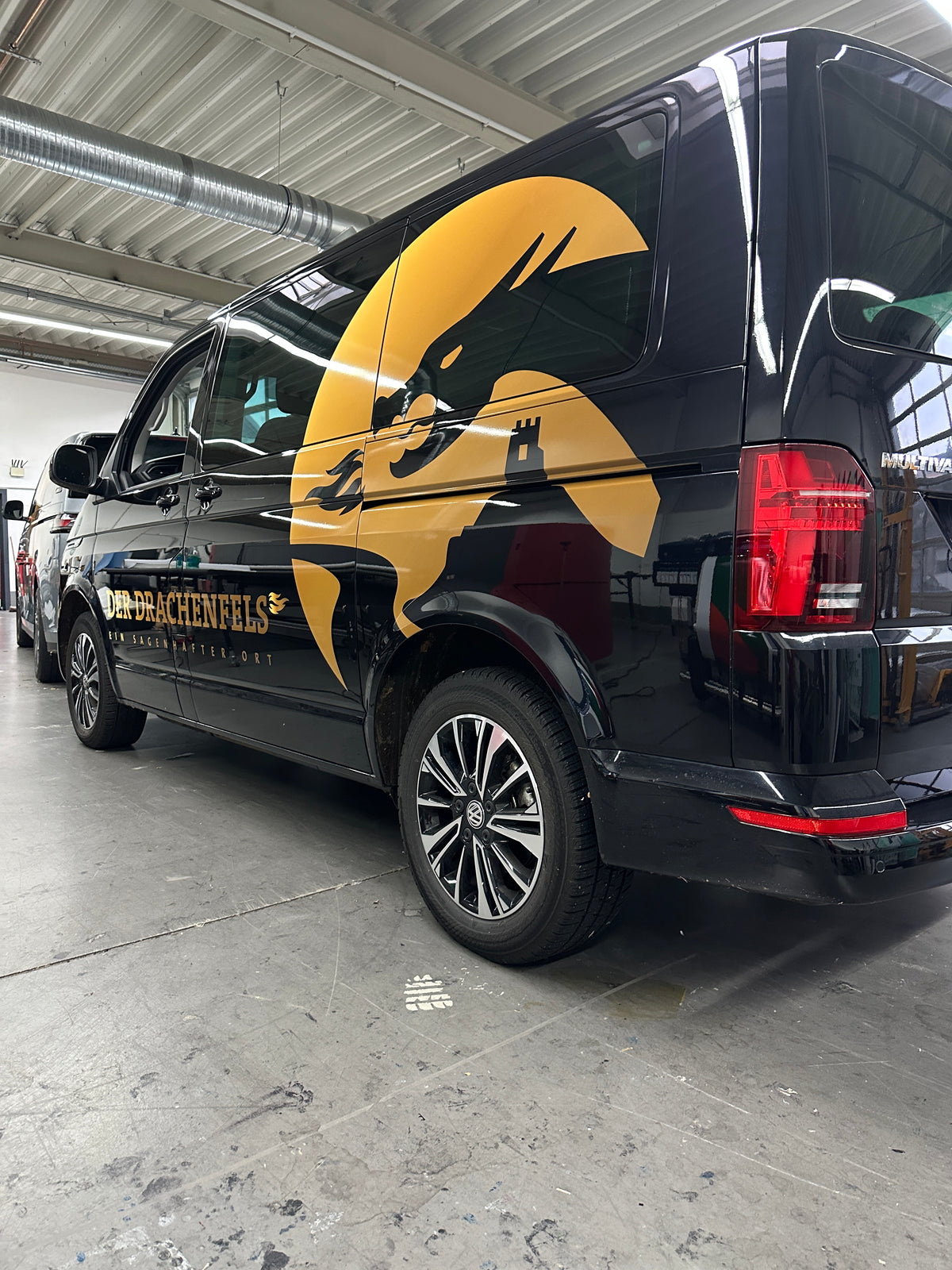 Fahrzeugbeschriftung eines schwarzen Transporters mit großer Drachenmotiv Teilfolierung und Logo Der Drachenfels auf der Fahrzeugseite.
