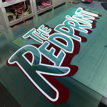 Weiß leuchtender LED Neon Schriftzug „THE REDPRINT“ auf türkisfarbenem und rotem Acrylglas in einer Werkstatt.