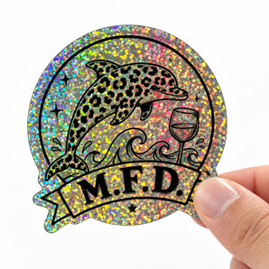 Eine Hand hält einen individuell gedruckten, holografischen Freiform-Sticker. Das schwarz gedruckte Motiv zeigt einen Delfin mit Leopardenmuster, Wellen, ein Weinglas und ein Banner mit der Aufschrift M.F.D. auf einer stark glitzernden Regenbogenfolie. Ein anschauliches Beispiel für Spezialeffekte, wenn man eigene Sticker drucken lässt.