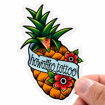 Eine Hand hält einen individuell konturgeschnittenen Aufkleber mit weißem Rand. Das Motiv im Traditional-Tattoo-Stil zeigt eine Ananas mit roten Blüten und einem hellblauen Banner, das den Schriftzug „hawaiiko_tattoo“ sowie ein kleines Instagram-Logo trägt. Ein passendes Beispiel für Künstler und Studios, die eigene Sticker drucken lassen, um sie als Werbemittel zu nutzen.