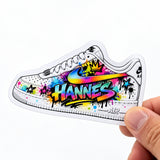 Eine Hand hält einen individuell gedruckten Freiform-Sticker mit einem weißen Rand. Der Aufkleber hat die Form eines Sneakers und ist mit farbenfrohen Graffiti-Spritzern, Sternen, einer Krone und dem zentrierten Schriftzug „HANNES“ verziert. Ein Beispiel für personalisierte Motive, die Kunden als eigene Sticker drucken lassen können.