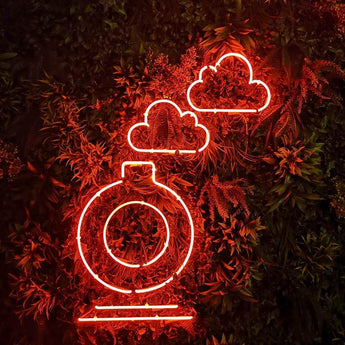 Orange leuchtendes LED Neon Schild in Form einer Flasche mit Wolken auf einer dekorativen, grünen Pflanzenwand.