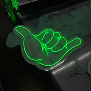 Grün leuchtendes LED Neon Schild in Form einer Shaka-Hand auf Acrylglas. Maßgeschneiderte Leuchtwerbung für Events.