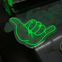 Grün leuchtendes LED Neon Schild in Form einer Shaka-Hand auf Acrylglas. Maßgeschneiderte Leuchtwerbung für Events.