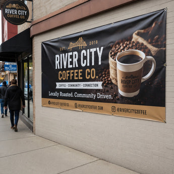 PVC Werbebanner der „River City Coffee Co.“, das direkt an einer Außenhauswand befestigt ist. Das Banner zeigt Kaffeebohnen und eine Tasse Kaffee und dient als hochwertige Außenwerbung für das lokal ansässige Café. Die Befestigung erfolgt über Metallösen am Rand des Banners.