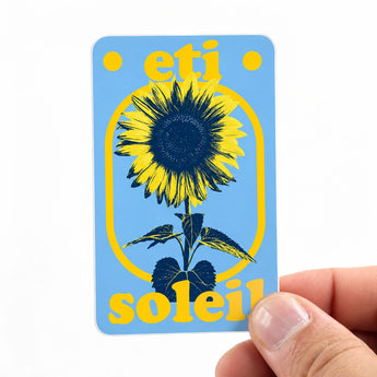 Eine Hand hält einen rechteckigen Aufkleber mit abgerundeten Ecken. Das Design im Retro-Stil zeigt eine stilisierte Sonnenblume in Gelb und Dunkelblau auf einem hellblauen Hintergrund, eingefasst von einem gelben Bogen. Ergänzt wird das Motiv durch die gelben Schriftzüge „eti“ oben und „soleil“ unten. Ein inspirierendes Beispiel für farbenfrohe Kunstwerke, die man als individuelle Sticker drucken lassen kann.