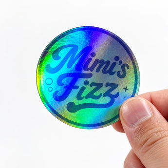 ine Hand hält einen runden, stark schillernden holografischen Aufkleber, der in Blau-, Grün- und Violetttönen leuchtet. Auf dem Aufkleber steht in dunkelblauer Retro-Schreibschrift „Mimi's Fizz“, ergänzt durch kleine gezeichnete Luftbläschen und Sterne. Ein anschauliches Beispiel für auffällige Materialeffekte, wenn man eigene Sticker drucken lässt.