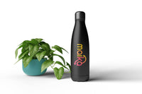 Thermobecher bedrucken – Eine matte, schwarze Isolierflasche mit dem farbenfrohen „maika“ Logo im Farbverlauf neben einer grünen Zimmerpflanze im türkisen Topf auf weißem Grund.