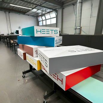 Mehrere große, rechteckige Produktdummys aus bedruckter Wellpappe, die in einer Produktionshalle auf einem schwarzen Rollwagen gestapelt sind. Die 3D-Aufsteller sind in verschiedenen Farben wie Blau, Rot, Gelb, Grau und Türkis bedruckt und tragen groß das Logo „HEETS“.