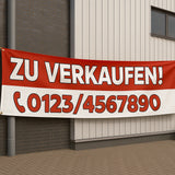 Ein rot-weißes "Zu Verkaufen" Werbebanner, befestigt an einer modernen Gebäudefassade. Passende Roll-Up Banner drucken.